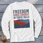 White long-sleeve t-shirt with red and blue graphic text reading "FREEDOM RINGS LOUDEST IN THESE HILLS" above layered mountain silhouettes.