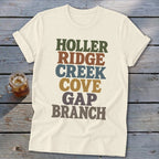 Cream-colored t-shirt with retro multicolored text reading HOLLER RIDGE CREEK COVE GAP BRANCH laid flat on wood.