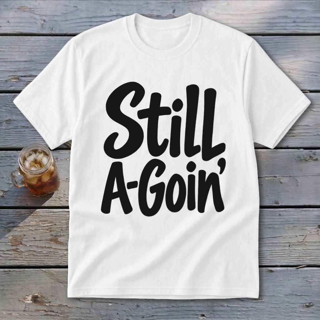 A white short-sleeve t-shirt with the black text Still A-Goin centered on the chest, lying on a wooden surface beside a glass of iced tea.
