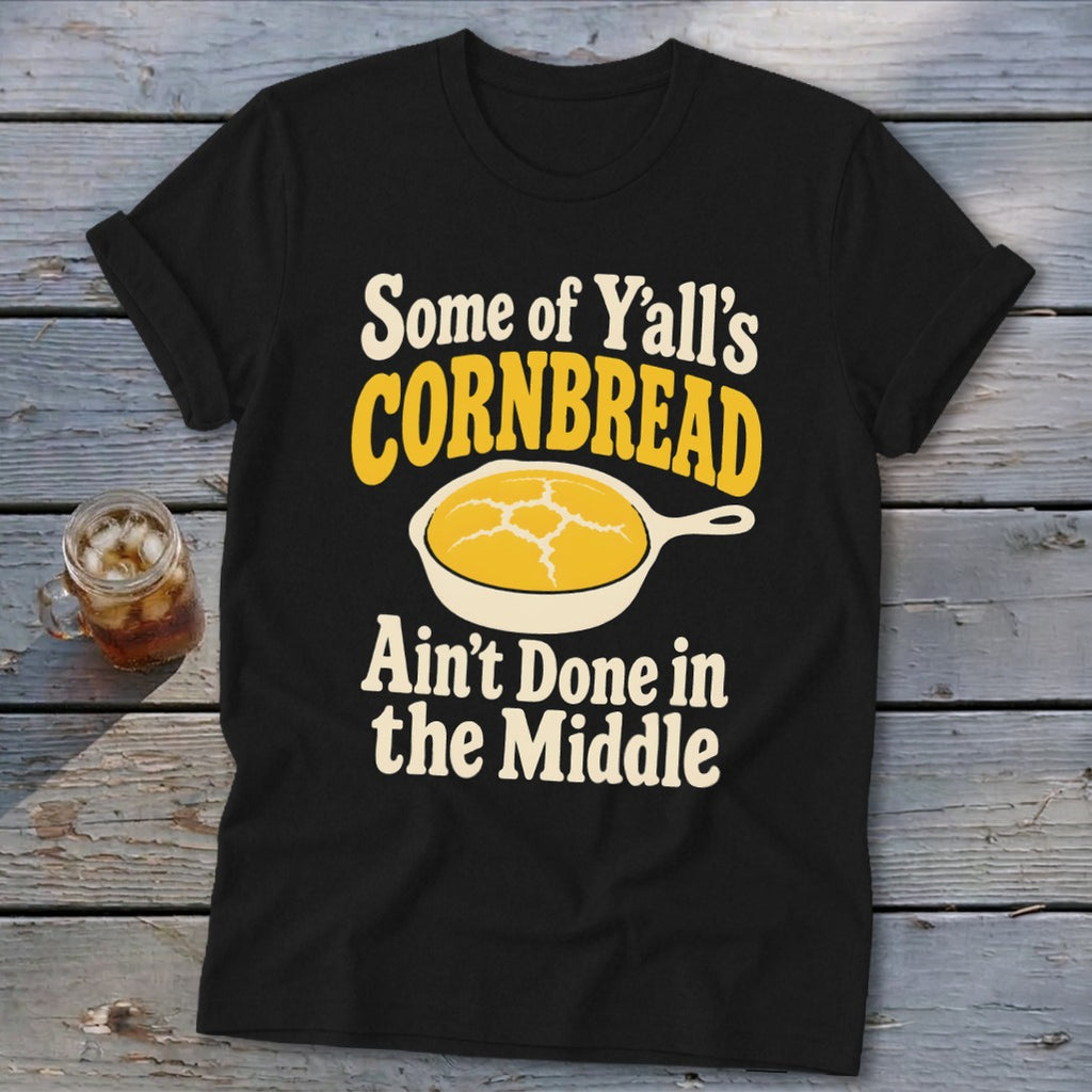 A black t-shirt with a graphic of cornbread in a skillet and text that reads Some of Y'all's Cornbread Ain't Done in the Middle.