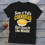 A black t-shirt with a graphic of cornbread in a skillet and text that reads Some of Y'all's Cornbread Ain't Done in the Middle.