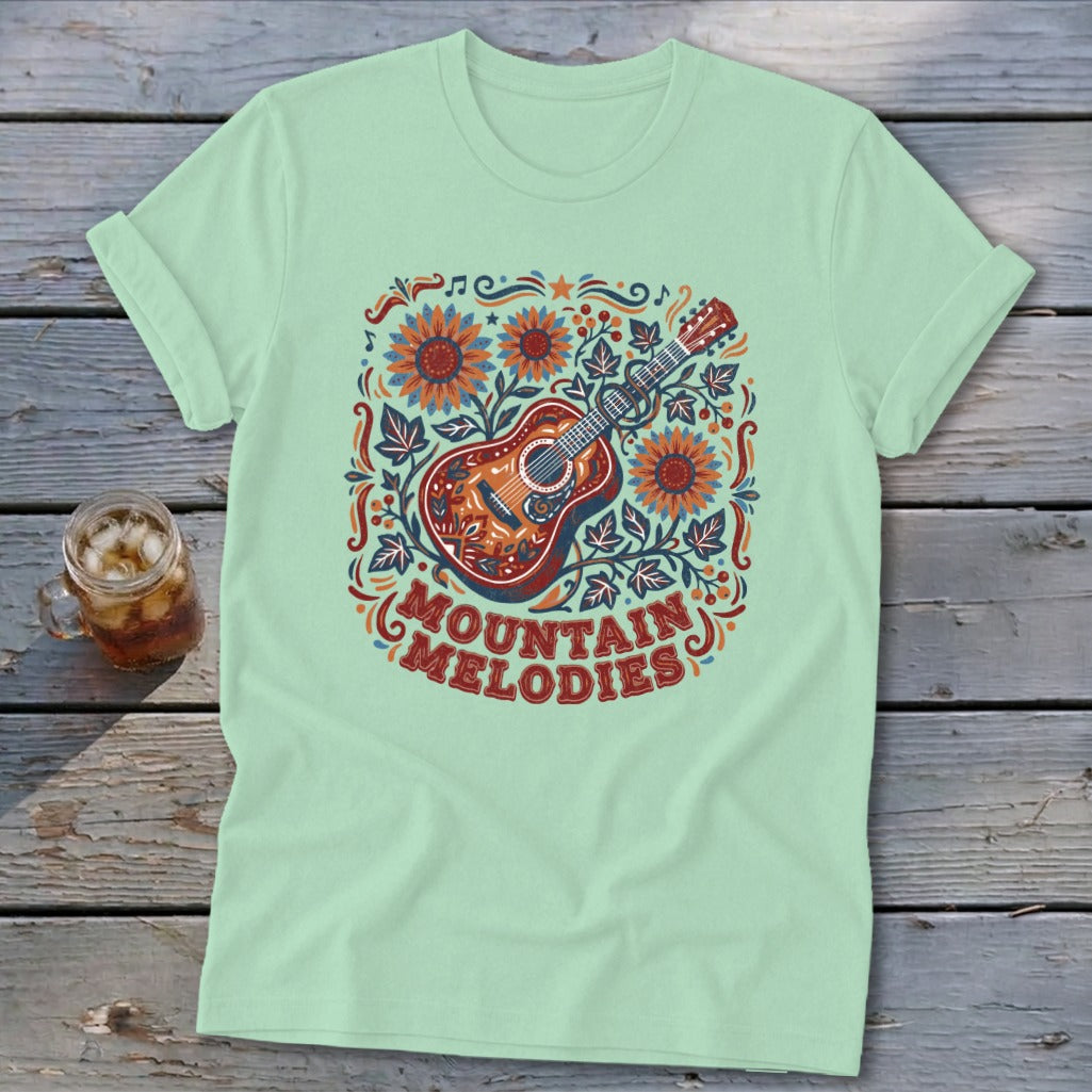 A light green t-shirt with a vintage graphic featuring an acoustic guitar, sunflowers, and the text MOUNTAIN MELODIES.