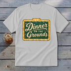 Light gray t-shirt with a marquee-style sign graphic reading Dinner on the Grounds, laid on a wooden surface.