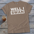 Brown t-shirt featuring the white text WELL I SWAN lying on a weathered wooden surface next to a glass of iced tea.