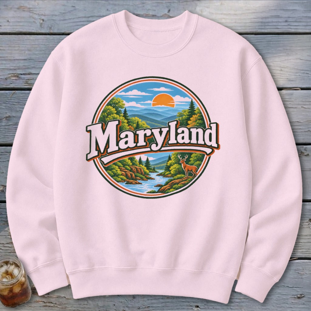 Pink sweatshirt with a circular nature graphic featuring "Maryland" text, a river, deer, and mountains.