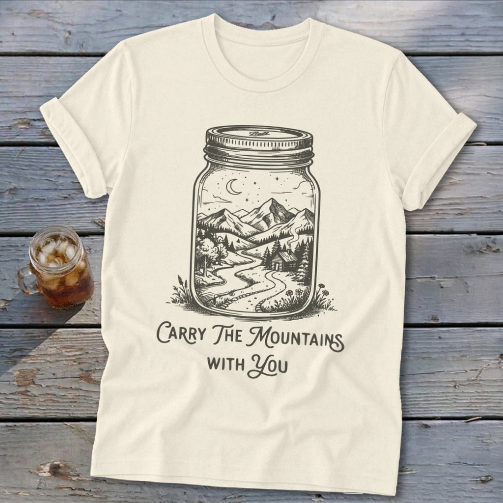 An off-white t-shirt featuring a black line art graphic of a landscape inside a mason jar, with the text "Carry The Mountains With You."