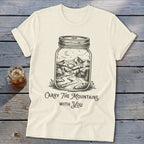 An off-white t-shirt featuring a black line art graphic of a landscape inside a mason jar, with the text "Carry The Mountains With You."