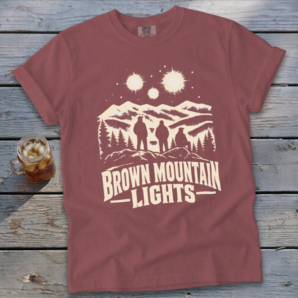 A mauve crewneck t-shirt featuring a graphic of three figures, mountains, and the text Brown Mountain Lights.