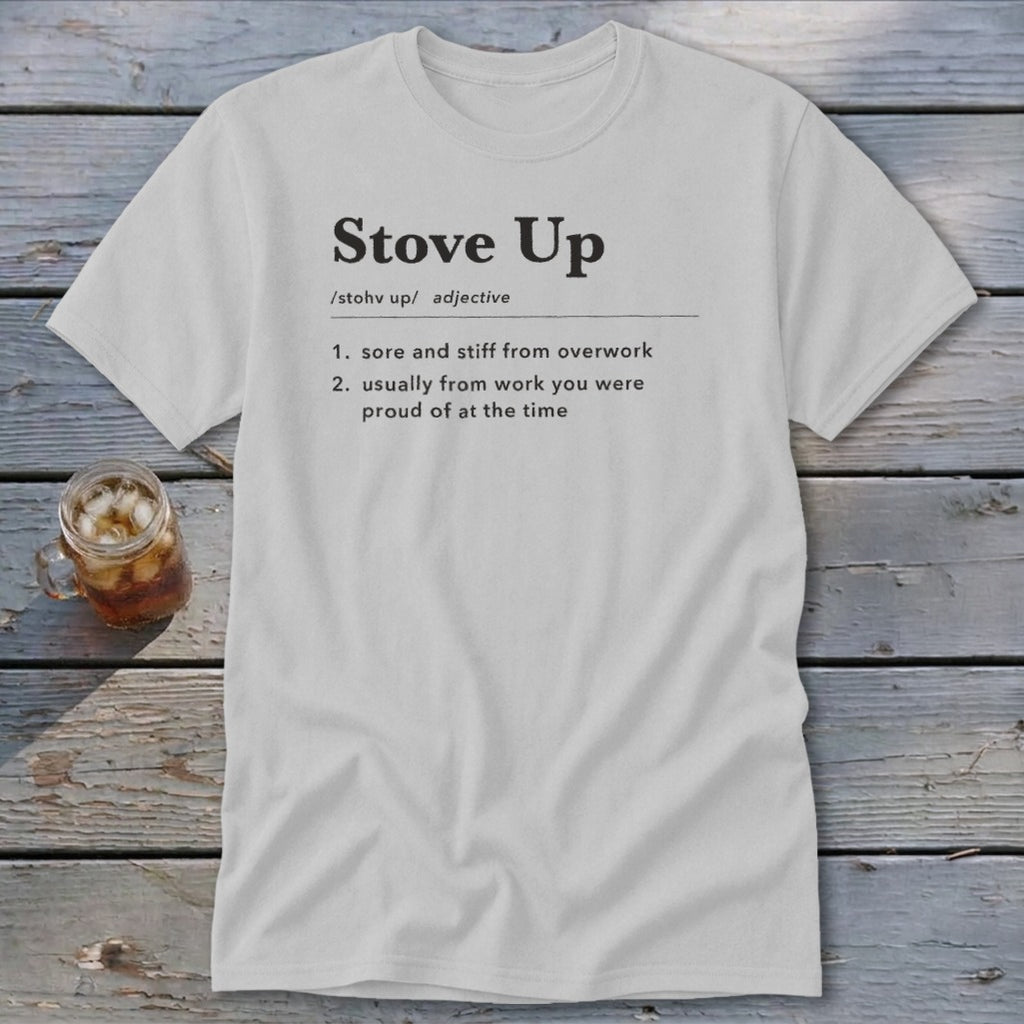 A light gray t-shirt with the definition of Stove Up printed on the chest, lying on a rustic wooden surface with a glass of iced tea nearby.