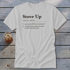 A light gray t-shirt with the definition of Stove Up printed on the chest, lying on a rustic wooden surface with a glass of iced tea nearby.