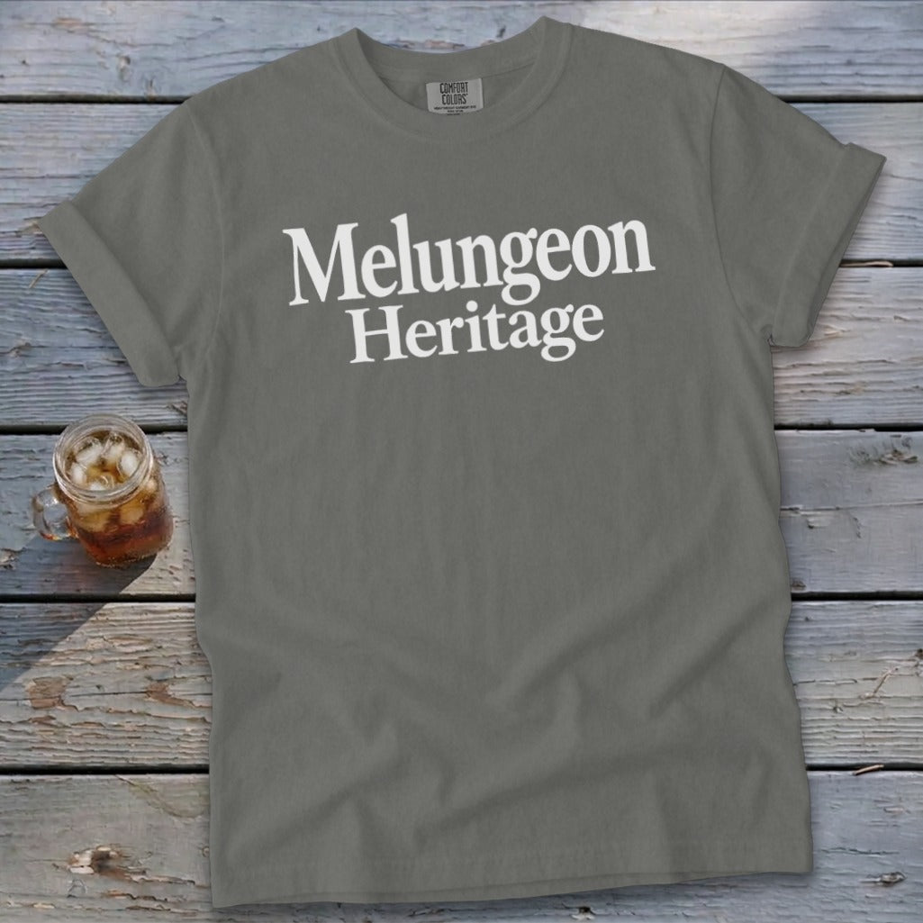 A gray t-shirt with rolled sleeves features the white text "Melungeon Heritage" on a wooden background.