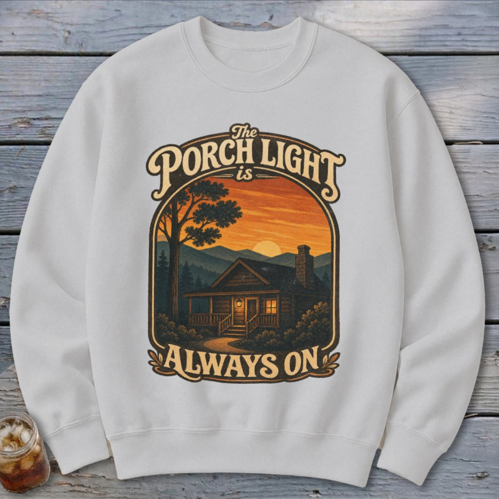 White sweatshirt with graphic: "The Porch Light is Always On" and cabin at sunset.