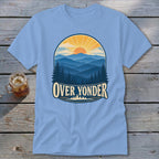 Light blue t-shirt featuring a mountain and sunrise graphic with the text Over Yonder, laid on a wooden surface.