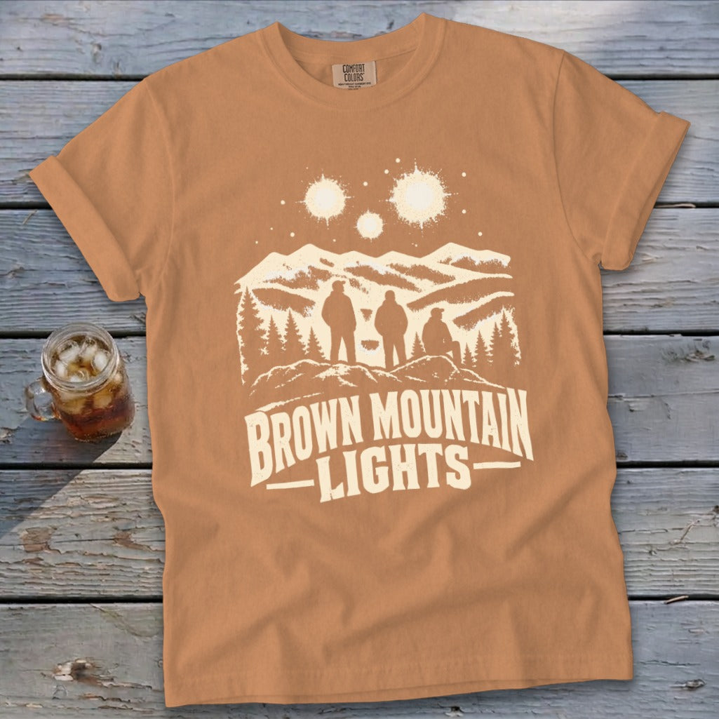 Tan t-shirt featuring a graphic of three figures looking at glowing orbs over mountains and the text "BROWN MOUNTAIN LIGHTS".