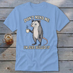 Light blue t-shirt with a grumpy opossum holding a coffee cup and text.