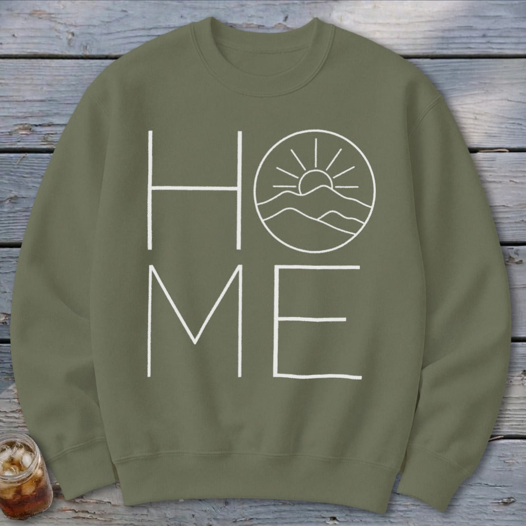An olive green crewneck sweatshirt featuring a white graphic that spells HOME with mountains and sun replacing the letter O.