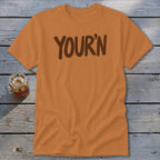 Orange t-shirt with brown "YOUR'N" text, displayed on wooden planks with a drink.