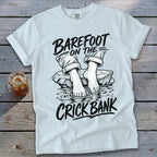 White t-shirt with black graphic of bare feet on rocks in water, text "BAREFOOT ON THE CRICK BANK".