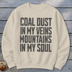 A cream-colored crewneck sweatshirt printed with the black text COAL DUST IN MY VEINS MOUNTAINS IN MY SOUL.