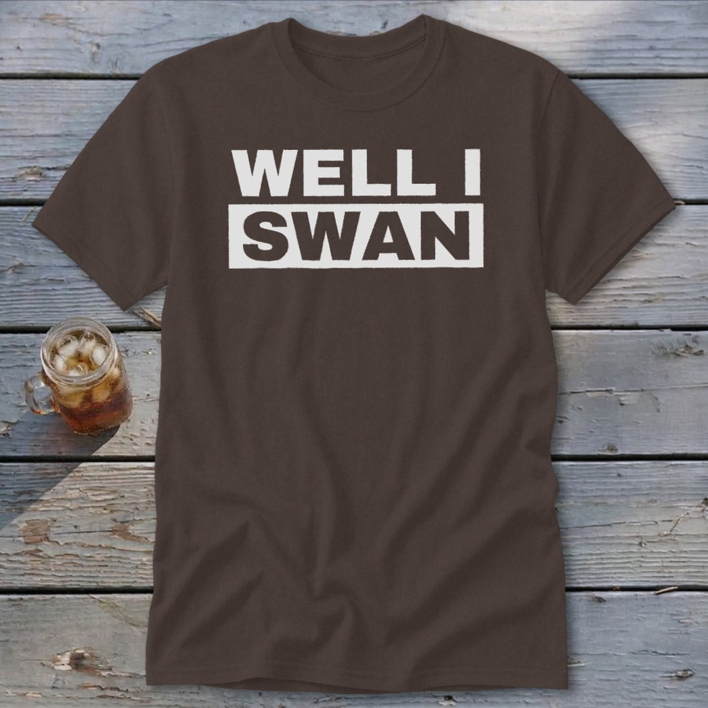 Brown crewneck t-shirt with the white text WELL I SWAN laid out on two lines, resting on a weathered wooden surface next to a glass of iced tea.