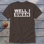 Brown crewneck t-shirt with the white text WELL I SWAN laid out on two lines, resting on a weathered wooden surface next to a glass of iced tea.