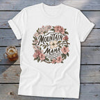 White t-shirt with "Mountain Mama Raised Em Right" text inside a floral wreath.