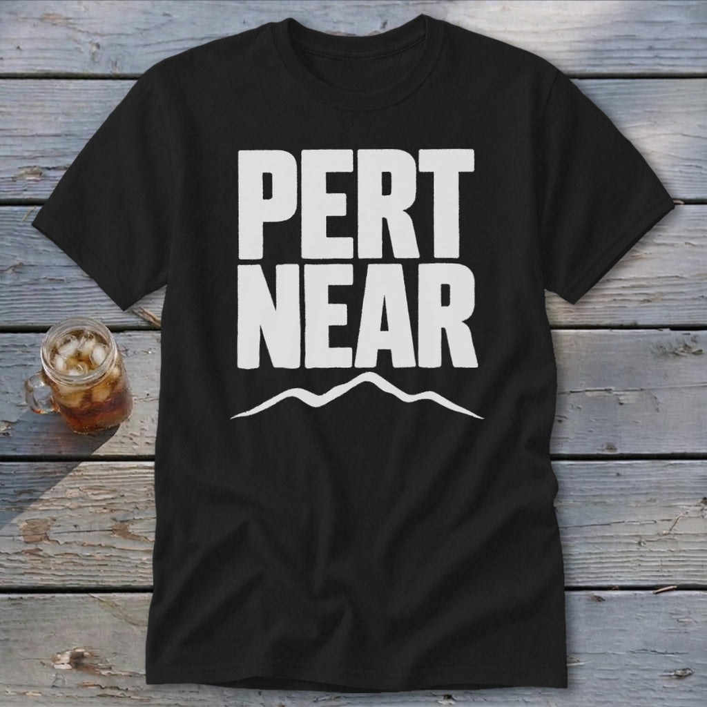 A black short-sleeved t-shirt with white block text reading PERT NEAR above a mountain outline, laid on a wooden surface beside a glass of iced tea.