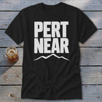 A black short-sleeved t-shirt with white block text reading PERT NEAR above a mountain outline, laid on a wooden surface beside a glass of iced tea.
