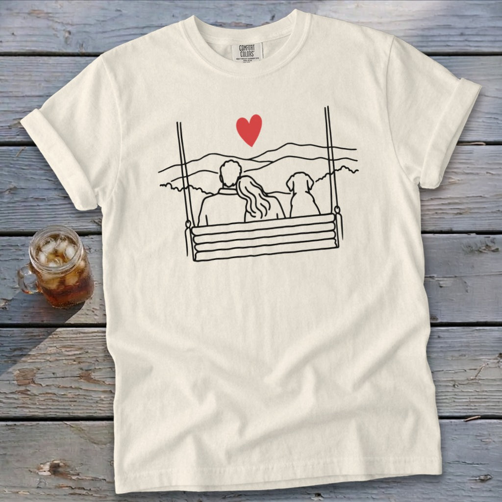 Line art of couple and dog on swing with heart and mountains, on cream t-shirt.