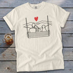 Line art of couple and dog on swing with heart and mountains, on cream t-shirt.