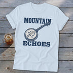 Light blue t-shirt featuring a banjo illustration with the text Mountain Echoes on a wooden surface.