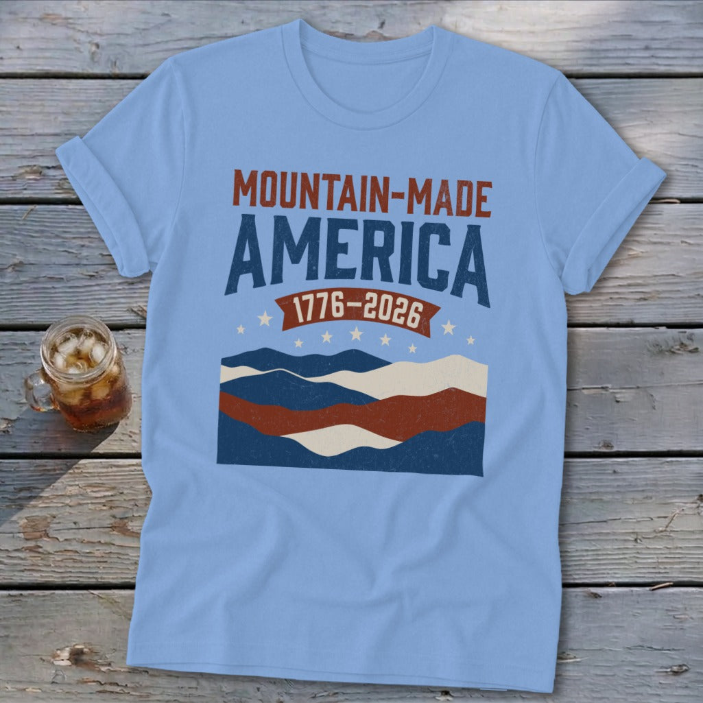 A light blue t-shirt printed with Mountain-Made America 1776-2026 text and wavy patriotic graphics.