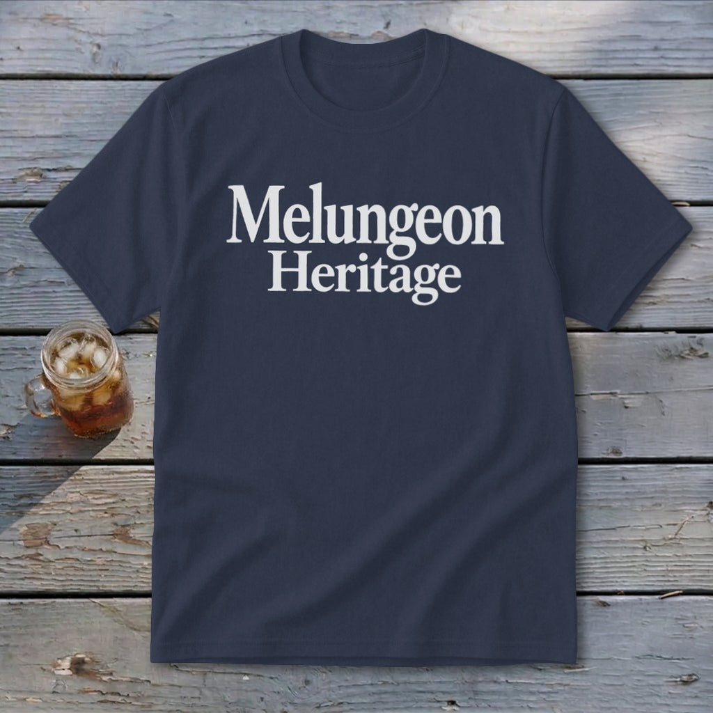 A dark blue t-shirt with white text reading Melungeon Heritage rests on a wooden surface beside a glass of iced tea.