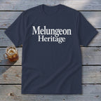 A dark blue t-shirt with white text reading Melungeon Heritage rests on a wooden surface beside a glass of iced tea.