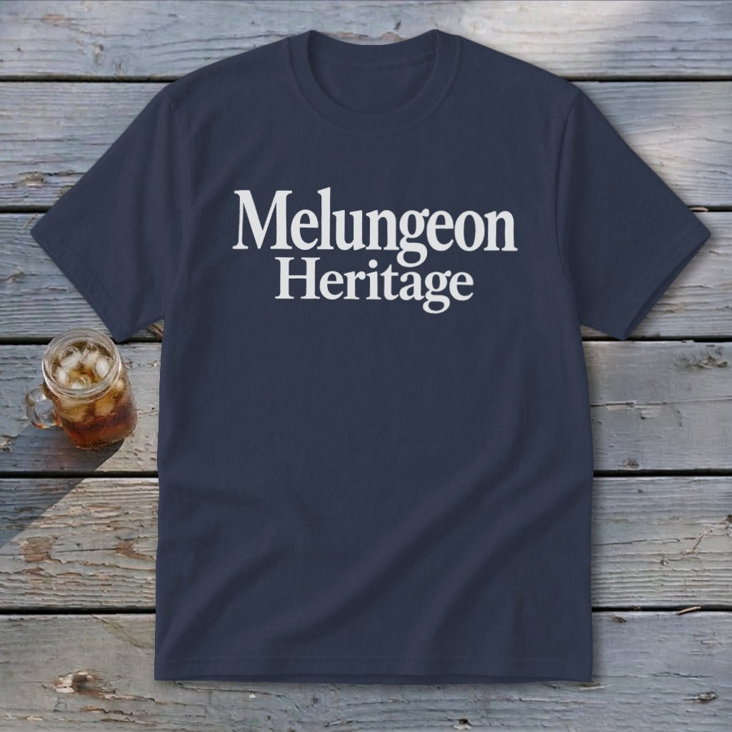 A dark blue t-shirt with white text reading Melungeon Heritage rests on a wooden surface beside a glass of iced tea.