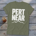 An olive green crewneck t-shirt with rolled sleeves lies flat on weathered grey wood, featuring large white block text reading "PERT NEAR" above a simple mountain outline.