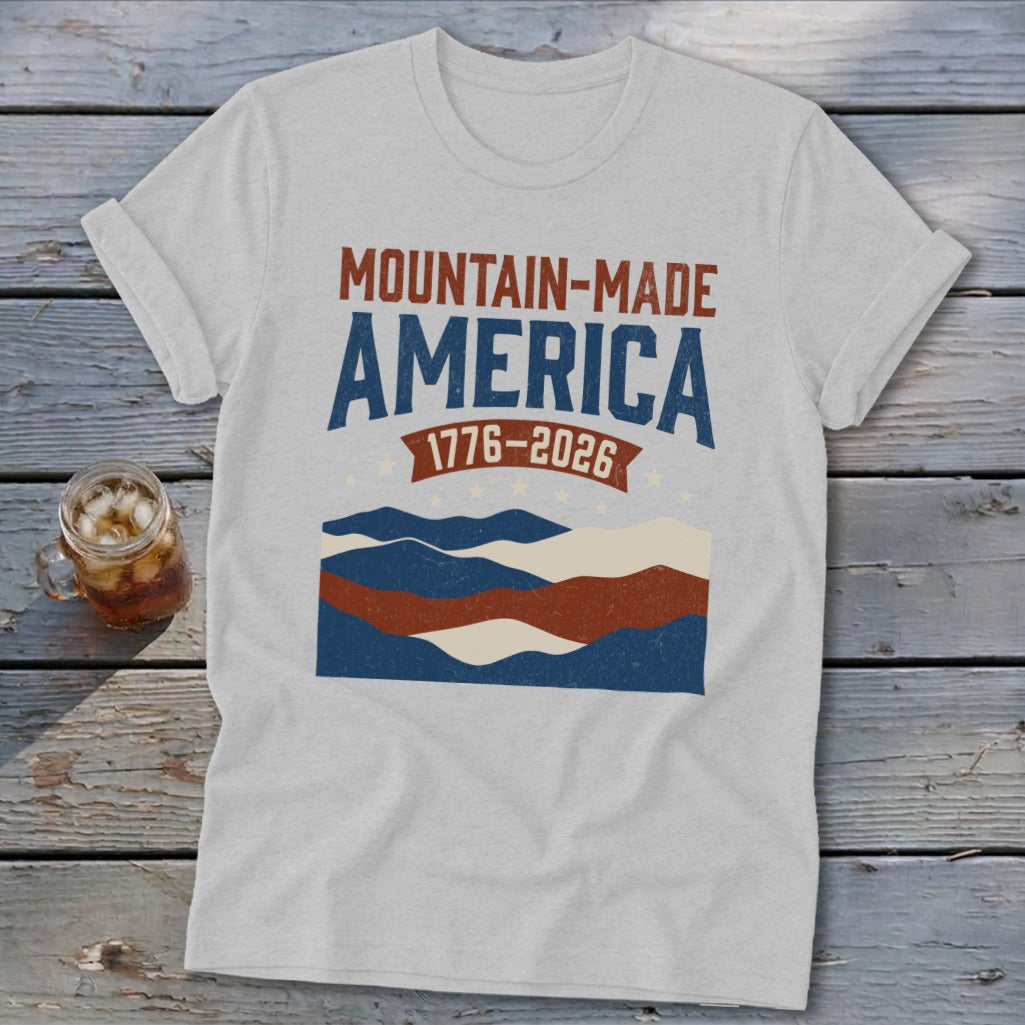 A grey t-shirt featuring a retro graphic with mountains and the text Mountain-Made America 1776-2026.