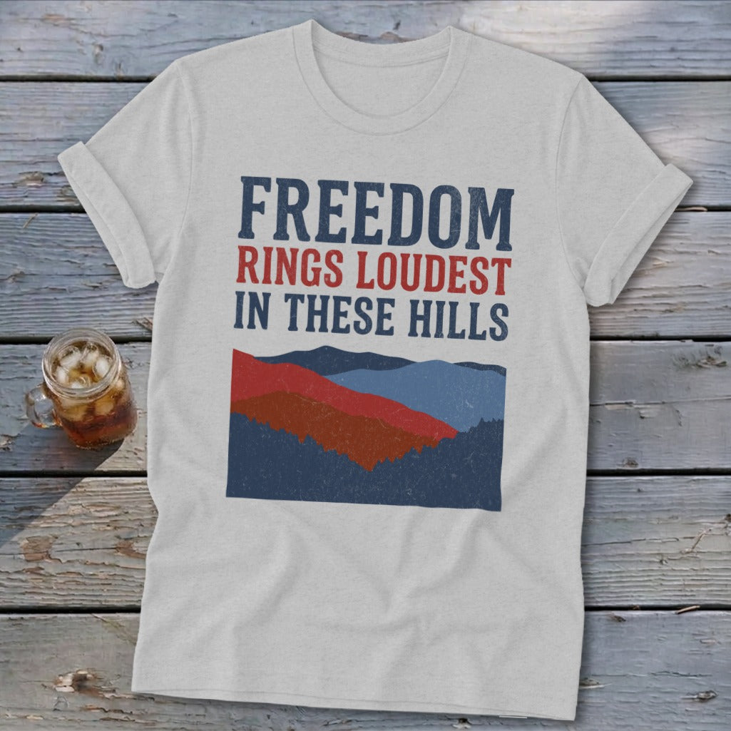 Light gray crewneck t-shirt featuring blue and red text reading FREEDOM RINGS LOUDEST IN THESE HILLS above a mountain graphic.