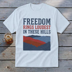 A white short-sleeve t-shirt features text reading Freedom Rings Loudest in These Hills above a mountain graphic.