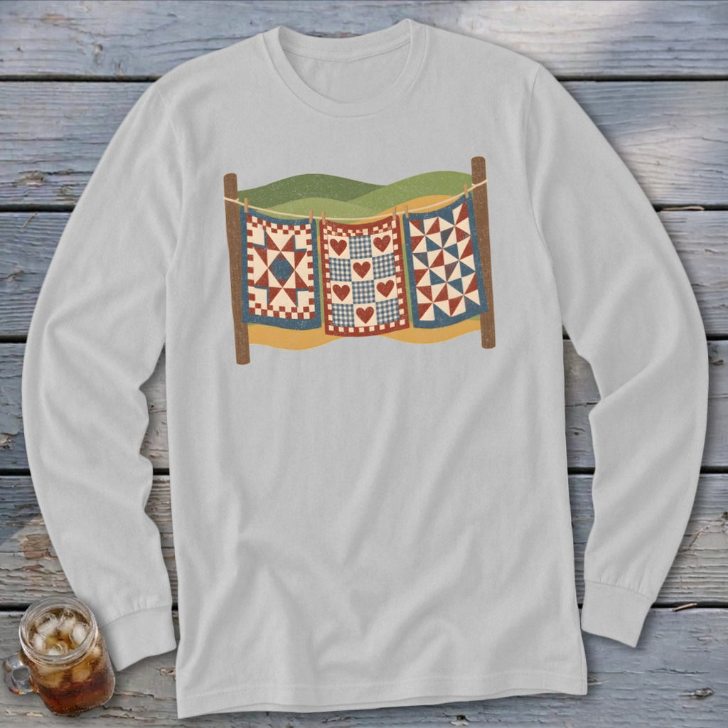 Light grey long-sleeve t-shirt printed with three vintage-style quilts hanging on a clothesline against rolling hills.