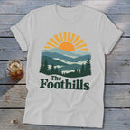 White t-shirt with "The Foothills" text and mountain sunset graphic.