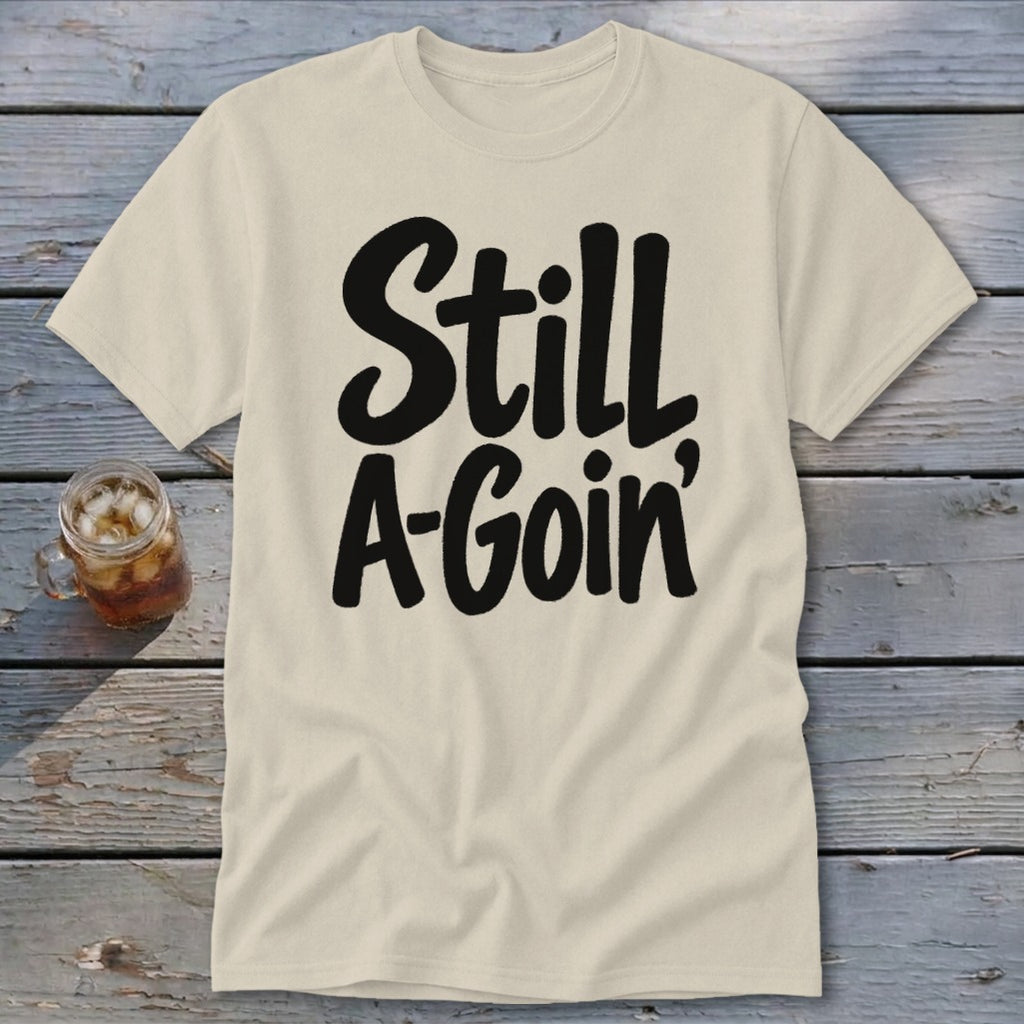 A cream-colored t-shirt with black cursive text reading Still A-Goin, displayed on a wooden surface beside a glass of iced tea.