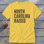 Yellow t-shirt with "NORTH CAROLINA RAISED" in dark gray text.