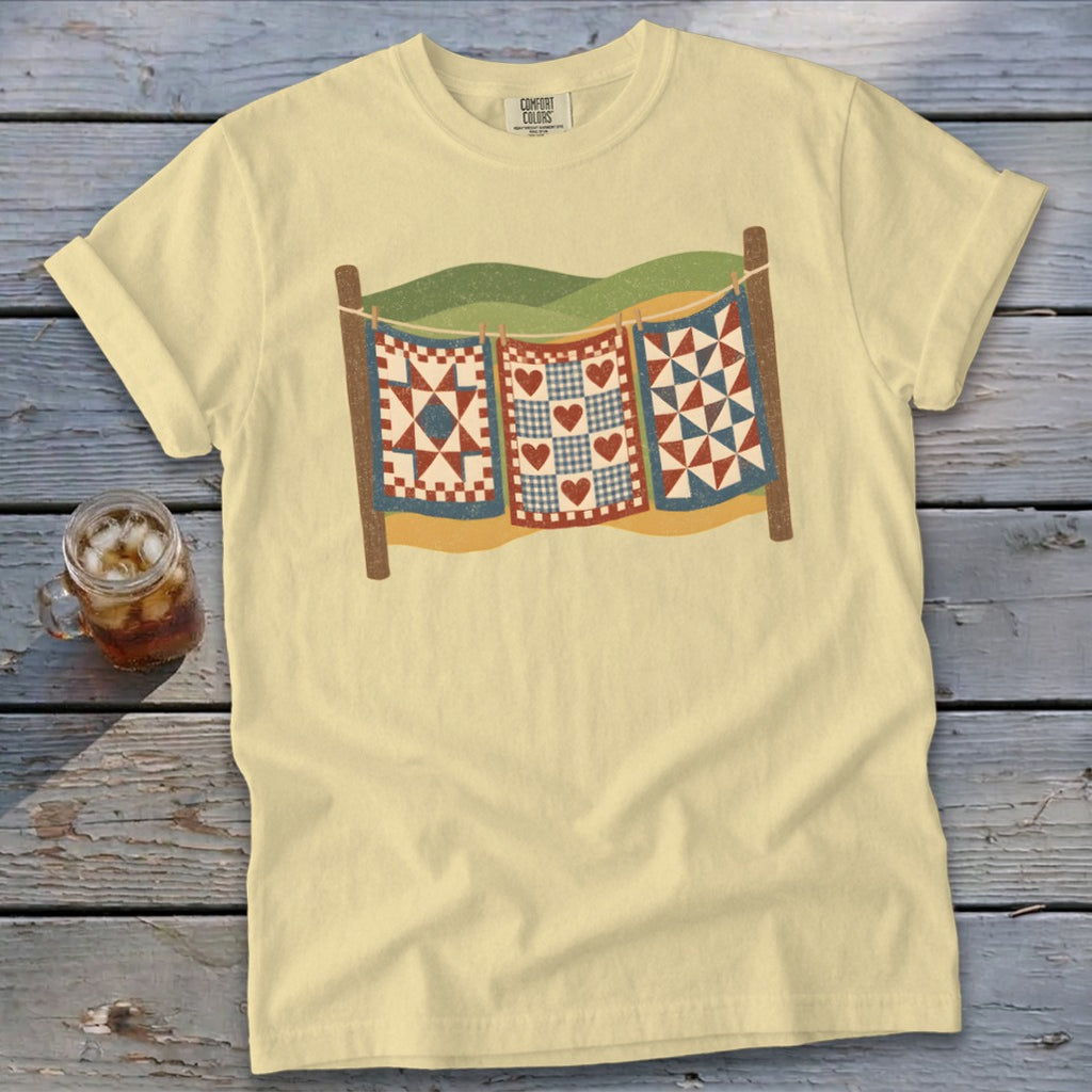 A pale yellow t-shirt featuring a graphic of three patchwork quilts hanging on a clothesline.
