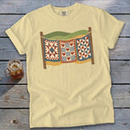 A pale yellow t-shirt featuring a graphic of three patchwork quilts hanging on a clothesline.