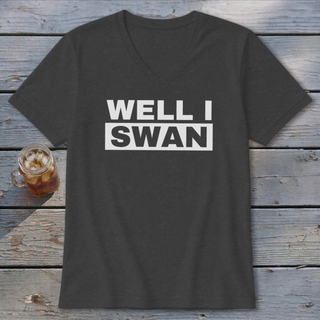 A dark gray v-neck t-shirt with white text reading WELL I SWAN on a wooden background.