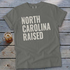 Gray t-shirt with "NORTH CAROLINA RAISED" text in white, on a wooden background.