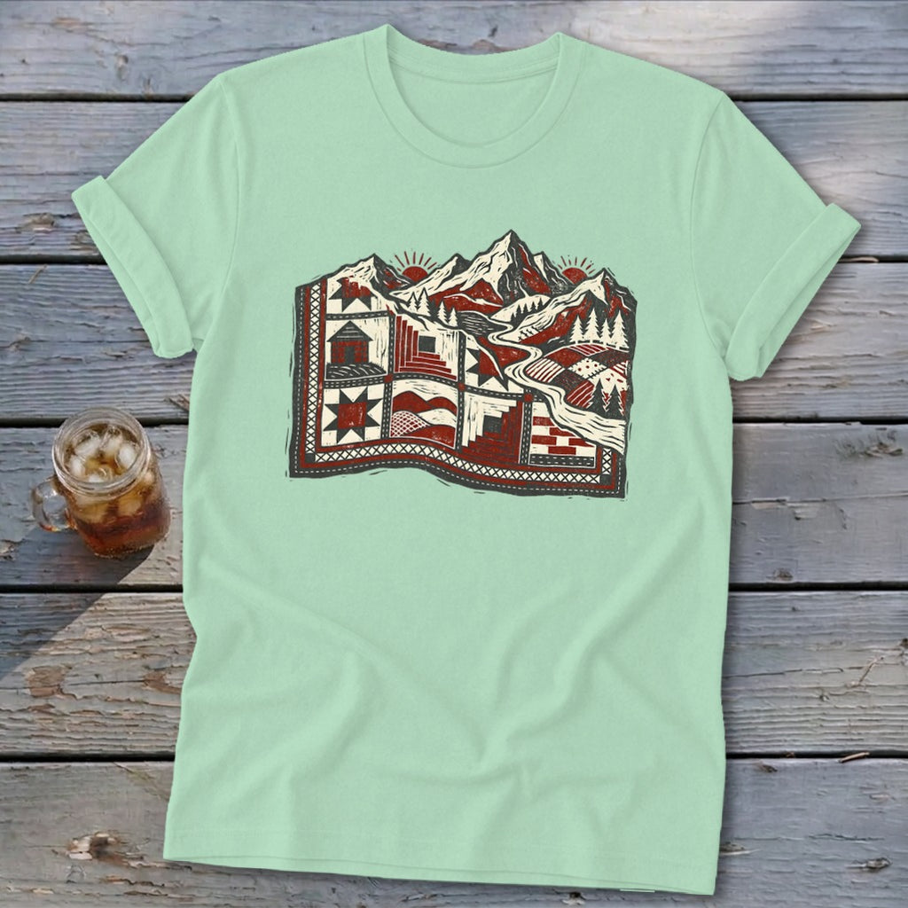 Light green t-shirt featuring a stylized graphic of a red and white patterned quilt transitioning into a snowy mountain landscape.