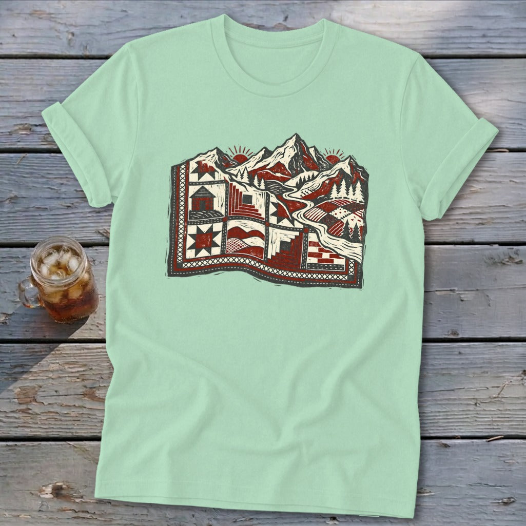 Light green t-shirt featuring a stylized graphic of a red and white patterned quilt transitioning into a snowy mountain landscape.