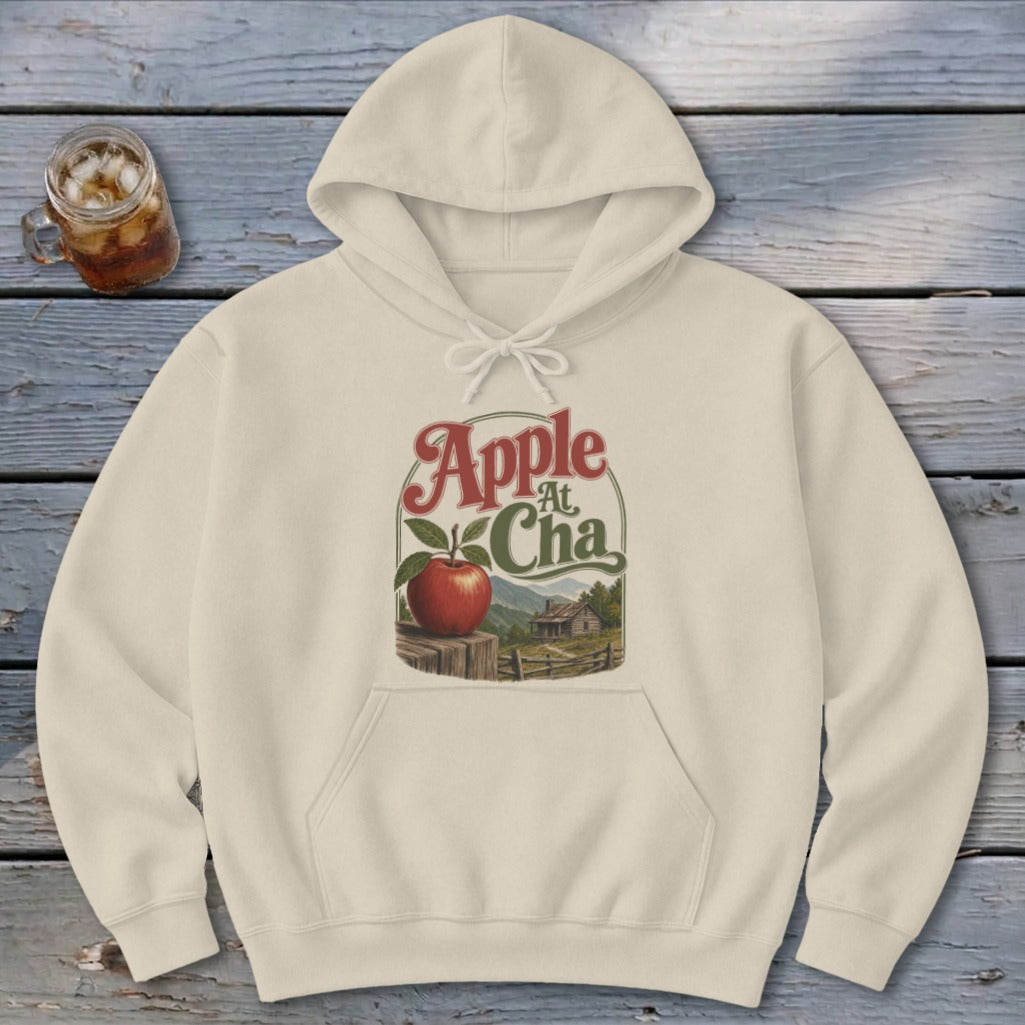 Cream-colored hooded sweatshirt featuring a graphic of an apple on a fence with mountains, a cabin, and Apple At Cha text.